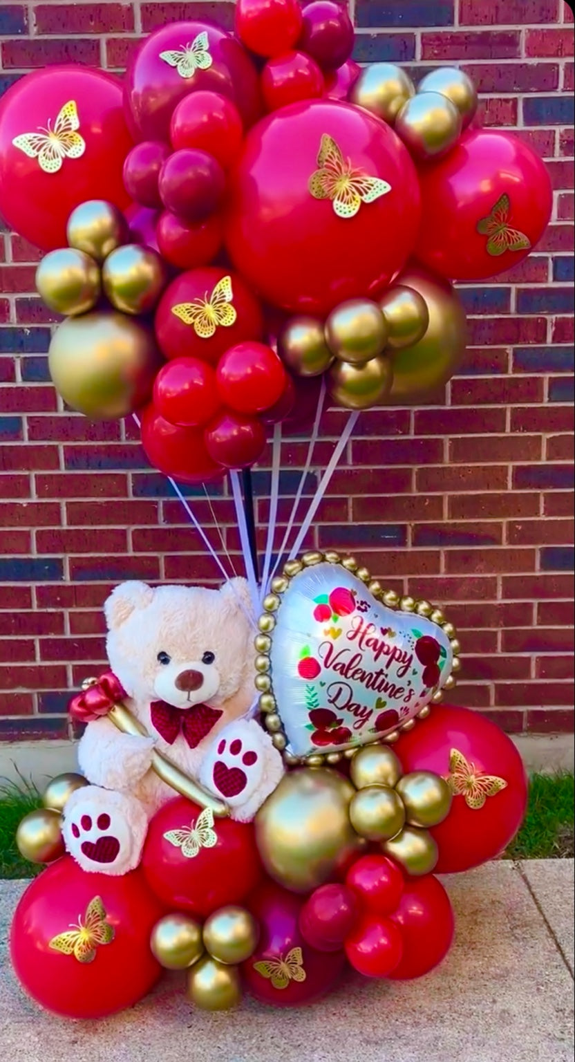 Valentine's Day balloon arrangement with a teddy bear and heart-shaped balloon against a brick wall.